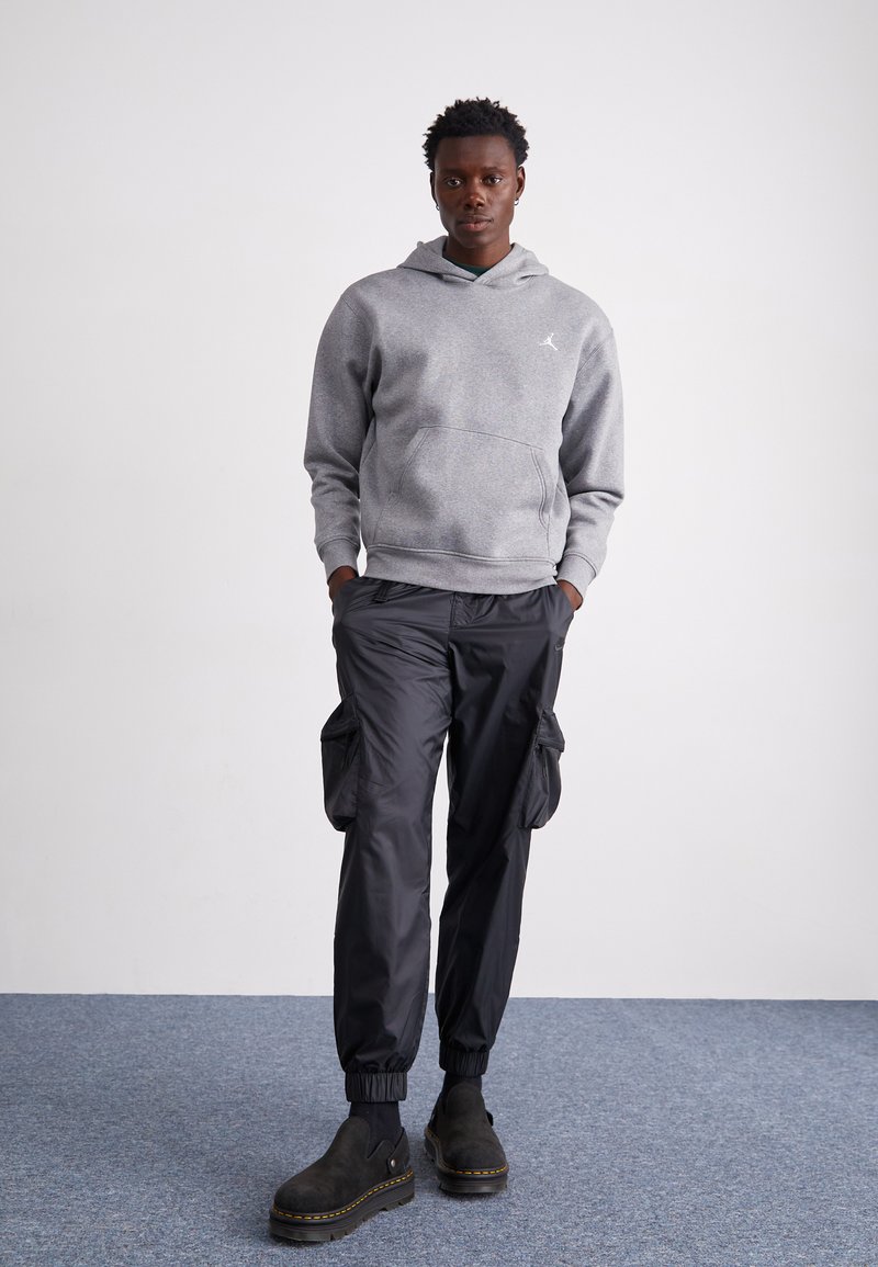 Grauer Kapuzenpullover mit Kängurutasche, schwarze Cargo-Hosen mit Reißverschlusstaschen und schwarze Knöchelstiefel vor einem blauen Teppich als Hintergrund.