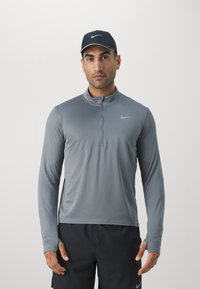 T-shirt de sport gris à manches longues avec col zippé, fabriqué dans un tissu lisse, arborant un logo Nike discret sur la poitrine. Casquette noire portée.