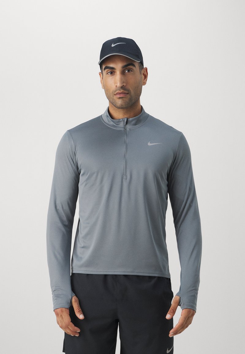 T-shirt de sport gris à manches longues avec col zippé, fabriqué dans un tissu lisse, arborant un logo Nike discret sur la poitrine. Casquette noire portée.