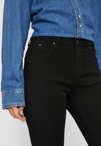 Μαύρα εφαρμοστά τζιν με λεία υφή και τυπικό σχέδιο πέντε τσέπες, σε συνδυασμό με ένα μπλε τζιν πουκάμισο με κλείσιμο με κουμπιά και contrasting μανίκια.