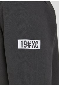 Tela de sudadera gris oscuro con una etiqueta rectangular blanca que lleva la inscripción "19#XC" en letras negras en negrita en la manga.