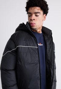 Sort polstret jakke med hætte; har hvide kanter langs sømmene og en frontlynlås. Bæres over en marineblå sweatshirt. Glat tekstur.