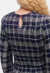 Femme blonde portant un haut à manches longues à carreaux bleu marine et blanc avec une fermeture bouton au dos en forme de trou de serrure et des détails smockés.