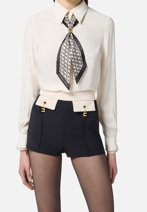 Mujer que lleva una blusa blanca de manga larga con un pañuelo de cuello con un estampado en blanco y negro y shorts negros de talle alto con bolsillos tipo solapa en beige y detalles dorados.