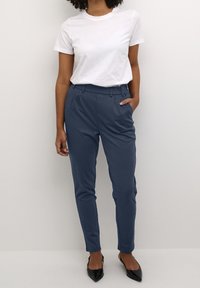 Femme portant une chemise blanche à manches courtes, un pantalon bleu ajusté avec les mains dans les poches, et des escarpins noirs à bout pointu, sur un fond uni.