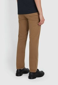 Chino marroni con vestibilità dritta, realizzati in un tessuto liscio. Presentano una vita e dettagli minimi. Indossati con scarpe nere.