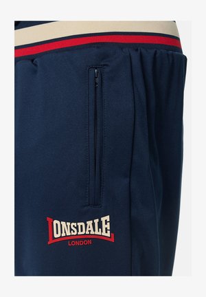 Mørkeblå joggers lavet af blødt stof med et off-white og rødt stribet taljebånd, med en lynlåslomme og et hvidt logo med røde detaljer.