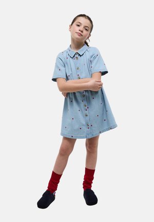 Jeune fille debout les bras croisés, portant une robe bleue à boutons ornée de petits cœurs rouges, des chaussettes rouges et des chaussures noires, sur fond blanc.