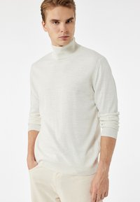 Suéter blanco de cuello alto con puños y dobladillo acanalados, hecho de una tela suave. El diseño presenta mangas largas y un ajuste relajado, mostrando una textura mínima.