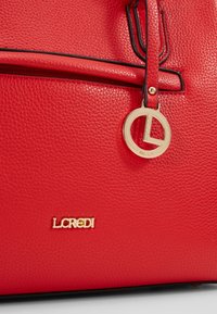 Sac à main rouge texturé avec accents noirs, arborant un logo doré et un charme circulaire. De plus, il possède une poche zippée à l'avant.