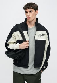 Αντιανεμικό μπουφάν Nike σε μαύρο και κρεμ, με φερμουάρ μπροστά, ελαστικές μανσέτες και λογότυπα. Φορεμένο πάνω από γκρι μπλουζάκι.