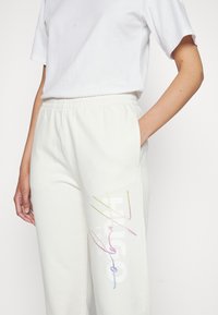 Krémové tepláky s elastickým pasem, ozdobené barevnými vyšívanými akcenty a logem potištěným na stehně.