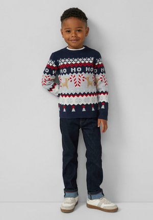 Jongen die glimlacht, gekleed in een marineblauwe vakantiesoftware met "HO HO HO", rendieren, zuurstokken, donkere spijkerbroek en witte sneakers, staande tegen een grijze achtergrond.