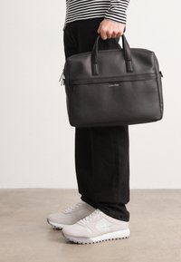 Briefcase nera in pelle con chiusura a zip, manici superiori, texture a grana sottile e un accent logo in metallo. Indossata con pantaloni neri e sneakers bianche.