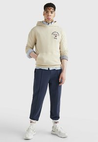 Felpa beige con logo e tasca, indossata sopra una camicia a quadretti azzurri chiari. Pantaloni tapered blu navy e sneakers bianche con dettagli rossi.
