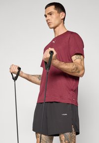 Bordeaux-rotes Sportshirt aus strukturiertem Stoff, kombiniert mit schwarzen Shorts. Hände halten schwarze Widerstandsbänder, die detaillierte Tattoos an den Armen zeigen.
