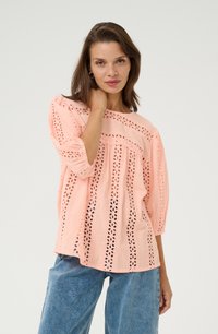 Blusa de color rosa claro con diseño de ojal, mangas 3/4 y corte holgado. La tela tiene una textura transpirable con recortes florales a lo largo del dobladillo.