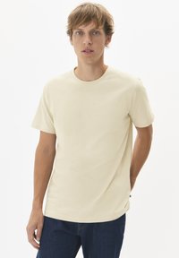 T-shirt beige clair à manches courtes avec un col rond et une texture lisse. Porté avec un jean en denim foncé, comportant une discrète étiquette logo sur le côté.