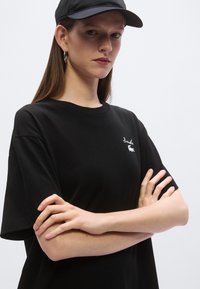 Čierne bavlnené tričko s krátkymi rukávmi a okrúhlym výstrihom, s bielym vyšívaným logom na hrudi. Bezšvové, s voľným strihom.