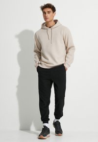 Sudadera beige clara con cordones, combinada con pantalones jogger negros. El modelo lleva zapatillas deportivas grises con suelas texturizadas y calcetines blancos.