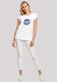 White cotton t-shirt with a round NASA logo in blue and red, paired with white slim-fit pants and white sneakers.