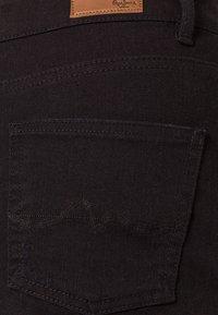 Pantalones vaqueros de mezclilla negra con un bolsillo trasero con costuras decorativas y una etiqueta de marca de cuero marrón. Textura suave con un ajuste ceñido.