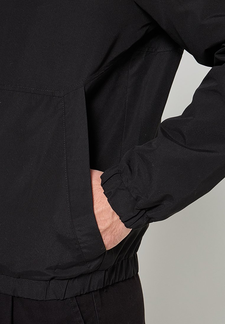 Veste coupe-vent noire avec des poignets élastiques, un ourlet resserré et des poches latérales. Le matériau semble léger et résistant à l'eau.