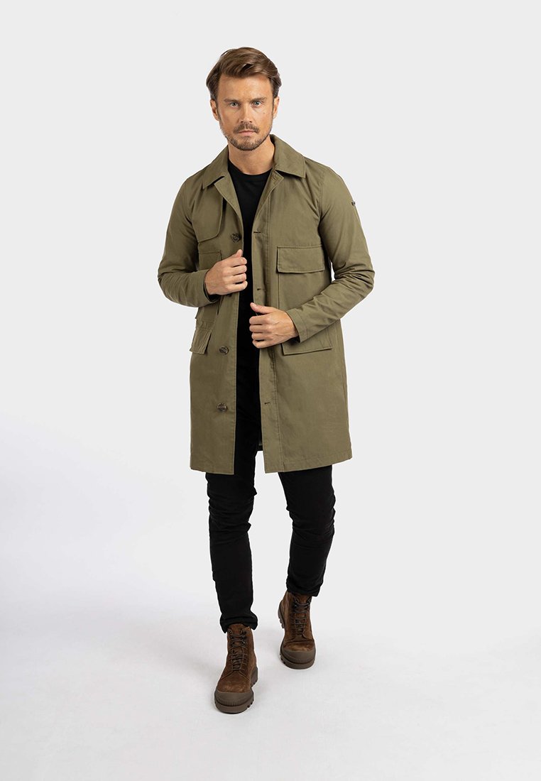 Trench coat verde oliva con chiusura a bottoni, due tasche sul petto, maniche lunghe e colletto classico, abbinato a pantaloni neri e stivali marroni.