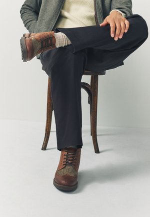 Mann sitzt auf einem Holzschemel mit einem übergeschlagenen Bein, trägt braune Leder- und Stoffschnürschuhe, dunkle Hosen und eine graue Jacke.