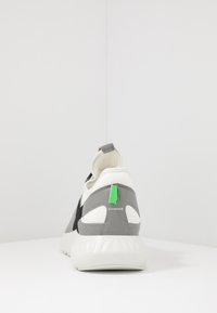 Baskets grises et blanches avec une tige en tissu, semelle en caoutchouc et un tirant vert. Présente des accents noirs et une forme de bout arrondi.