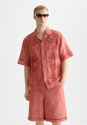 Uomo biondo con occhiali da sole scuri e camicia rossa a maniche corte con ricamo floreale, abbinata a pantaloncini rossi coordinati.
