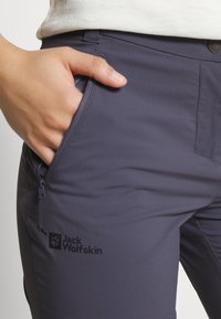 Tmavě šedé kalhoty Jack Wolfskin s hladkou texturou, boční kapsou na zip a logem vytištěným na stehně. Detailní pohled s rukou v kapse.