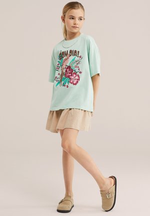 Joven con una camisa azul clara de gran tamaño con estampado de pájaros y flores, falda beige, zapatos sin cordones y un collar, de pie con una pierna hacia adelante.