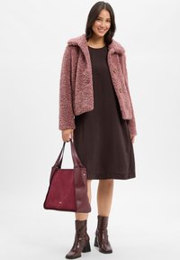 Frau in einer pinken, flauschigen Jacke über einem dunkelbraunen Kleid, die eine burgunderfarbene Handtasche hält und braune Ankle Boots mit Blockabsätzen trägt.