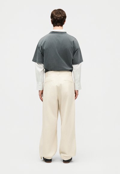 Homme debout de dos portant une chemise grise à manches courtes par-dessus une chemise blanche à manches longues et un pantalon ample beige avec des chaussures noires.