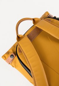 Gelbe Tasche mit einer glatten, matten Oberfläche, schwarzen Reißverschlüssen, Lederakzenten und gepolsterten Schultergurten für zusätzlichen Komfort.