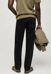 Vaqueros de denim negro con un diseño de pierna recta, combinados con una chaqueta beige. La persona sostiene la chaqueta con una mano y lleva sandalias marrones.