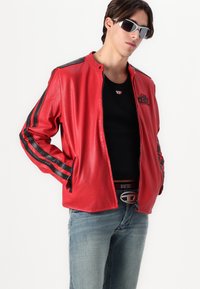 Raudona perforuota odinė striukė su juodomis juostelėmis, užtrauktuko priekyje ir stovinčiu apykakle. Dėvima per juodą marškinį ir mėlynus džinsus.