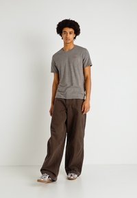 camiseta gris de manga corta con un pequeño logotipo, combinada con pantalones oscuros de corte holgado y zapatillas metálicas sobre un fondo sencillo.