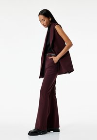 Bordeaux mouwloze blazer met een getailleerde pasvorm, in combinatie met bijpassende flared broeken en zwarte platform schoenen. Gladde stof en een minimalistisch design.
