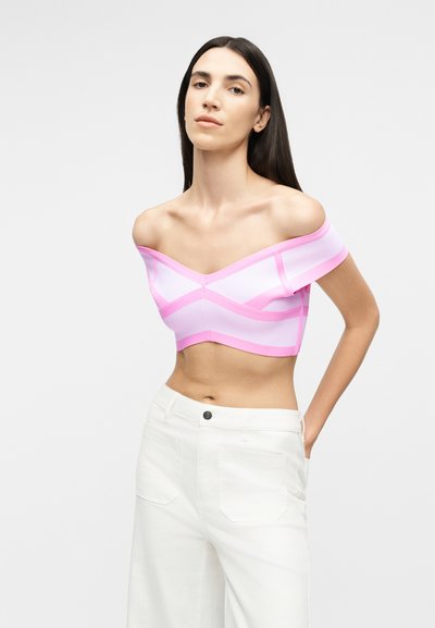 Γυναίκα με μακριά σκούρα μαλλιά φοράει ροζ και λευκό τοπ έξω από τον ώμο και λευκό παντελόνι με ψηλή μέση, στέκεται μπροστά σε απλό φόντο.