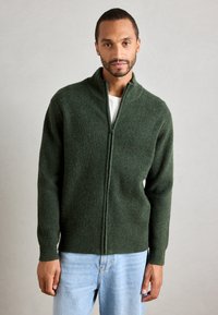 Mann iført en mørkegrønn jakke med glidelås over en hvit skjorte og lyseblå jeans, som står mot en ensfarget lys grå bakgrunn.