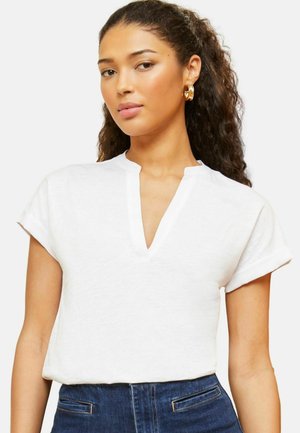 Kortsiklike juustega naine, kes kannab kuldseid rõngaskõrvarõngaid, valget lühikeste varrukatega V-kaelusega pluusi ja tumelõhnavat teksapükse ühevärvilise tausta ees.