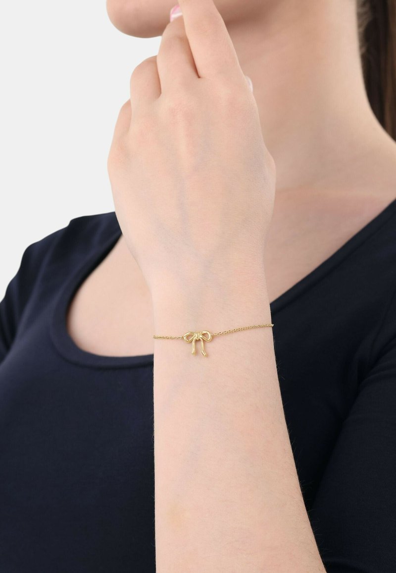 Pulsera de oro con un diseño de lazo sobre una delicada cadena, exhibida en una muñeca con un tono de piel suave, sobre un fondo liso.