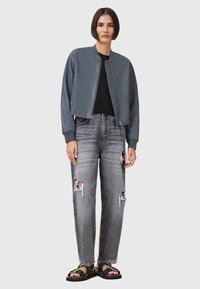 Giubbotto bomber in pelle grigia con zip frontale, abbinato a jeans grigi strappati con grandi buchi e sandali neri flat.