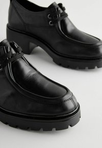 Schwarze Lederschuhe mit abgerundetem Zeh, klobigem Absatz und strukturiertem Profil; verfügen über eine Schnürung und erhöhte Nähdetails.