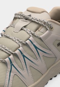 Μπεζ αθλητικό παπούτσι Columbia με κορδόνια με σχέδια, επάνω μέρος από διχτυωτό ύφασμα και μπλε ζιγκ-ζαγκ λεπτομέρειες στο πλάι, εμφανιζόμενο σε κοντινό πλάνο.