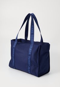 Sötétkék bevásárlótáska, két felső fogantyúval, textúrált anyagból készült. A pántokon kék "TOMMY JEANS" logóval rendelkezik. Téglalap alakú.