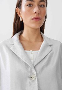 Silberner, leichter Blazer mit strukturiertem Finish, mit Revers und großen Knopfdetails, getragen über einem weißen, gerippten Oberteil mit Knöpfen.