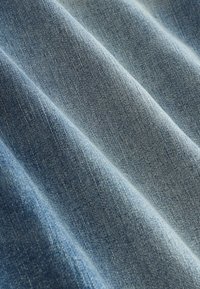 Denim tkanina v svetlom modrom odtieni, s jemnou štruktúrou a viditeľným tkaným vzorom, s miernym zvrásnením pre prirodzený, uvoľnený vzhľad.